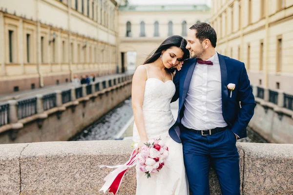 Gut aussehende junge Frau mit ansprechendem Aussehen trägt weißes Hochzeitskleid, hält schönen Strauß, lehnt sich an die Schulter des Liebhabers, genießt Zweisamkeit und ruhige Atmosphäre, als stünde sie allein auf der Brücke. — Stockfoto