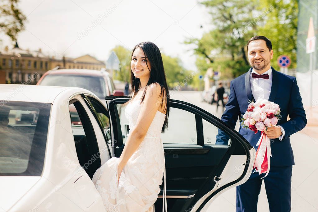 Portrait of newlywed couple sit in car after ceremony, have happy expression. Smiling female bride in white dress sits in white car, going to have cruise with bridegroom. Special occasion concept