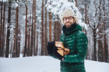 Unshaven smiling male holds pile of firewood, going to make fire to warm himself in frosty weather, spend winter day in forest, has picnic with friends outdoor. Fashionable unshaven man chop firewood clipart