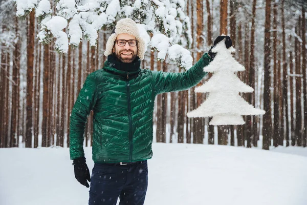 Look at this beautiful fir tree! Cheerful bearded man in fur cap with ear flaps and green anorak, glad to have walk in winter forest, enjoys beautiful trees and nature landscapes. Season concept — Stock Photo, Image