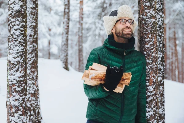 Outdoor Portret van glimlachen mannelijke staat in de buurt van boom bedekt met sneeuw, houdt van brandhout, ziet er gelukkig omhoog zoals aankondigingen eekhoorn op boom, nu in winter woud. Mensen en recreatie concept — Stockfoto