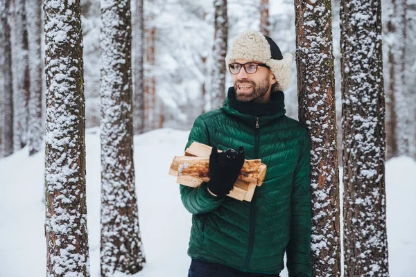 Gelukkig bebaarde man in groene jas, staat in de buurt van boom, houdt van brandhout, zorgvuldig opzij kijkt, heeft vrolijke expressie, tegen een achtergrond van besneeuwde bos. Modieuze peinzende man buiten — Stockfoto