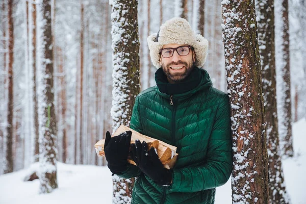 Good looking smiling male wears warm winter clothes holds firewood, stands near tree, spends free time with friends in winter forest, going to have picnic, makes fire to warm in frosty weather