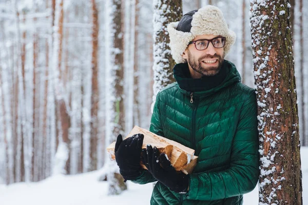 Bebaarde jonge man draagt een warme muts en groene jas houdt stapel brandhout, gelukkig opzij kijkt, merkt iets, houdt van winter en ijzig weer. Dreamful aantrekkelijke man besteedt tijd in bos — Stockfoto