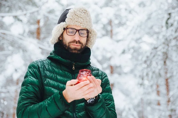 Attractive serious male wears warm winter clothes, keeps candle, stands against winter forest background, enjoys fresh air, being very confident. Fashionable unshaven man outdoors. — Stock Photo, Image