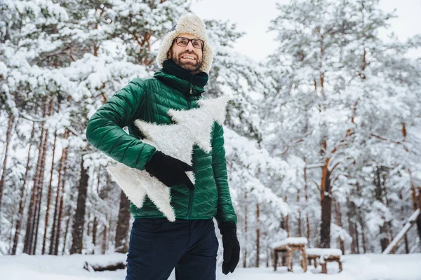 Glada manlig modell iklädd vinterkläder, innehar vit konstgjord fir tree, står i vinter skog, har trevligt leende, åtnjuter vackra landskap. Människor, helgdagar, rekreation koncept — Stockfoto