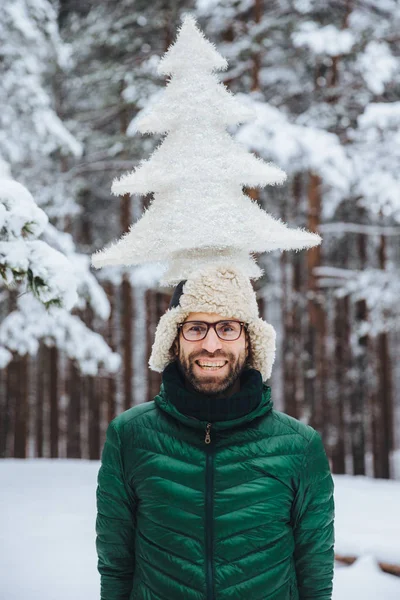 Vertical portrait of cheerful bearded man has fun alone in winter forest, keeps artificial fir tree, poses outdoors, admires frosty snowy weather, expresses positiveness and pleasant emotions — Stock Photo, Image