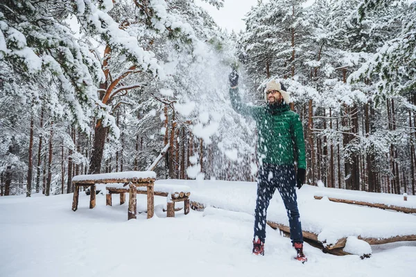 Buiten schot van knappe bebaarde man gekleed in warme kleding, heeft plezier als gooit sneeuw in lucht, vakantie doorbrengt in winter bos, spreekt positiveness. Recreatie en weer concept — Stockfoto