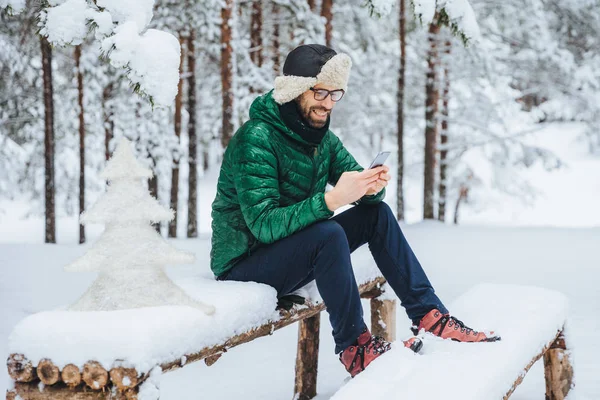 Neşeli erkek iletişim akıllı telefon ve yalnızlık, sakin atmosferi sahiptir, kış orman, yakın arkadaşı ileti almak için mutlu kar ile kaplı tahta bankta oturuyor — Stok fotoğraf