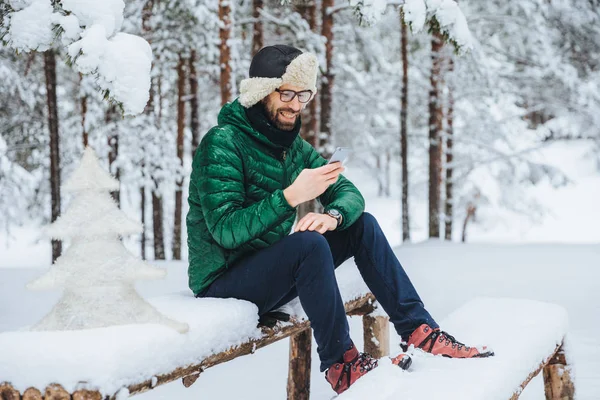 Glimlachend positieve male warme winter kleding draagt, bericht op mobiele telefoon leest, besteedt vrije tijd in de rustige sfeer buiten in de winter bos, geniet van frisse lucht. Mensen, rust, technologie concept — Stockfoto