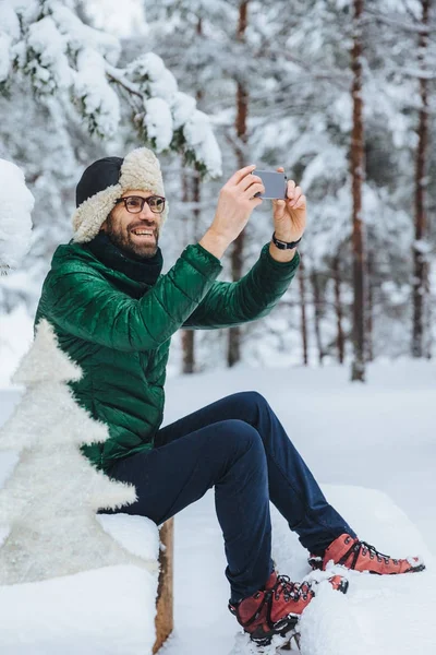 Verticale portret van aangename op zoek mannelijke maakt foto met slimme telefoon maakt opnamen van mooie winterlandschappen, geniet van natuur- en frozy weer, heeft aangename glimlach. Mensen, technologie concept — Stockfoto