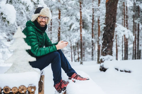 Photo of cheerful male types messages on smart phone looks directly into camera, sits in white winter forest, smiles happily, enjoys communication, watches video, admires landscapes