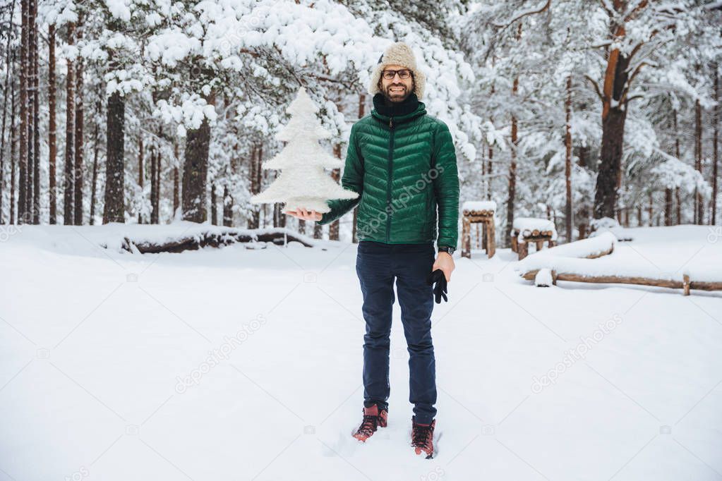 Outdoor shot of good looking male wears warm hat and green jacket holds winter fir tree, poses against trees covered with snow, looks happily directlly into camera. Recreation and season cpncept