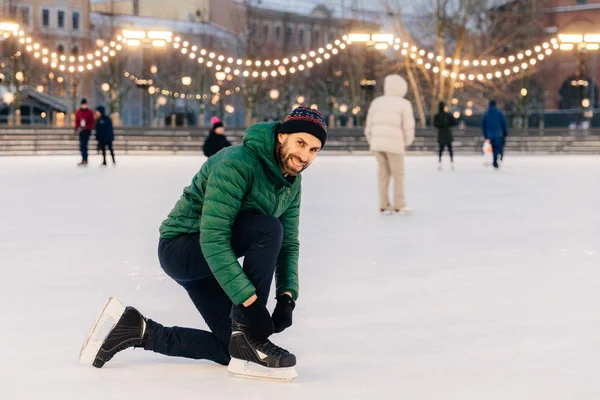 Outdoor shot of man in green coat, laces up skates, looks happil — Stock Photo, Image