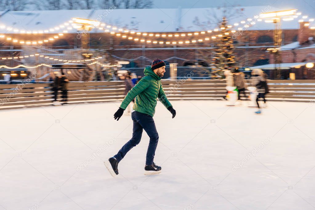 Pleased man in green jacket, wears skates, goes skating on ice, 