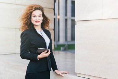 Sevimli kadın freelancer lüks hafif saçlı, mavi gözlü, kırmızı dudaklar ve saf deri, siyah resmi takım elbise, üstünde onun iş ortakları ile toplantı olacak elektronik aygıtı n el ele tutuşarak kızarmış