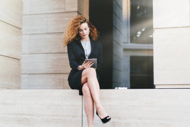Şık takım ve yüksek topuklu ayakkabılar, iletişim için modern tablet kullanarak ince bacakları olan kıvırcık saçlı genç kadın modeli. Dışında çalışan, ciddi görünümlü genç serbest meslek