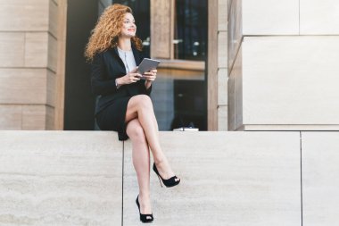 Portre siyah takım elbise ve modern tablet ile oturan ince uzun bacakları olan yüksek topuklu ayakkabılar içinde zarif güzel kadın bir kenara mutlu bir ifadeyle bakıyor. Açık poz işçisi kadın ofis