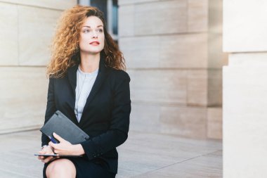 Gür saçlı, mavi gözlü, kırmızı dudaklar, yakışıklı genç iş kadını tutan eller cep kitabı ve modern tablet bilgisayar, siyah takım elbise giyen bir kenara düşünceli ifade ile seyir