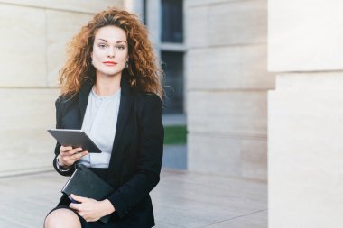 Zarif Bayan Beyaz bluz, siyah ceket ve etek giymiş office builduing yanında otururken tablet ve cep kitabı tutarak portresi. Kıvırcık saçlı, güzel yüzünü başarılı iş kadını