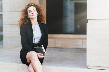 Çok güzel kadın modeli gür dalgalı saçlı, kırmızı dudaklar ve parlayan gözleri, resmen tablet cep kitabı, güvenle kameraya bakarak ile tutarak bacaklarını geçerken giyinmiş. İş kavramı