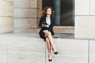 Kadın girişimci ince bacakları ile oturan siyah resmi kostüm giyen holding modern dijital tablet bilgisayar, ona gelecekte ortaklık planlama düşünceli ifade ile seyir bacak bacak