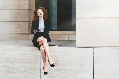 Sevimli kıvırcık yalnız oturan kendine güvenen bakışla ince uzun bacaklarını gösteren etek, siyah ceketli zamanlamaları veya diyagramları, iş raporu yapma arıyor tablet, holding giyimli kadın
