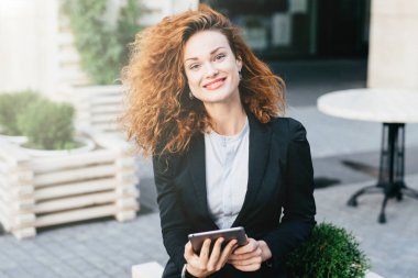 Siyah takım elbise ve tablet ile açık kafede oturan forması giyen genç bayan kıvırcık saçlı, kırmızı boyalı dudaklar ve parlayan gözleri, hoş kameraya gülümseyen. Teras, çekici Bayan