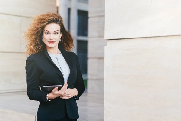 Young successful businesswoman in elegant clothes, having luxuriant hair, holding tablet in hands while going for work. Gorgeous female holding digital tablet computer. Technology and business — Stock Photo, Image