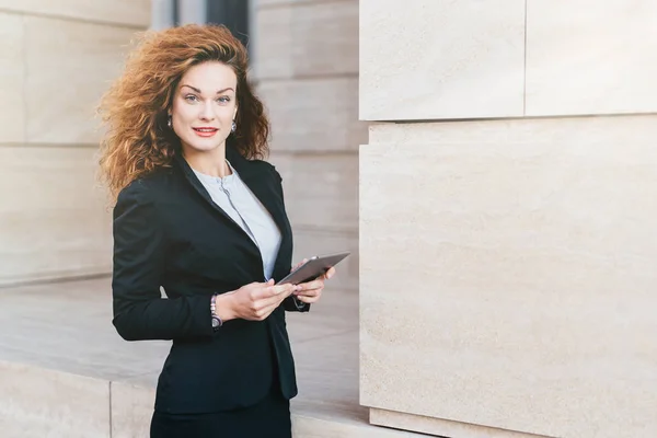 Mulher de negócios jovem elegante bonita em traje preto, tendo cabelo encaracolado e aparência atraente, segurando tablet, lendo notícias na Internet enquanto posando contra a grande acumulação na rua — Fotografia de Stock