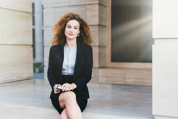 Attractive young woman wearing black formal suit, sitting crossed legs with electronic device waiting for business partner near cafe. Gorgeous female with appealing appearance posing at camera — Stock Photo, Image