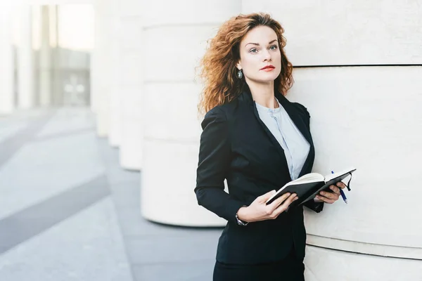 Pretty young businesswoman wearing black jacket, skirt and white blouse, holding her pocket book with pen, writing notes or looking for spare days in order to arrange meeting with companions — Stock Photo, Image