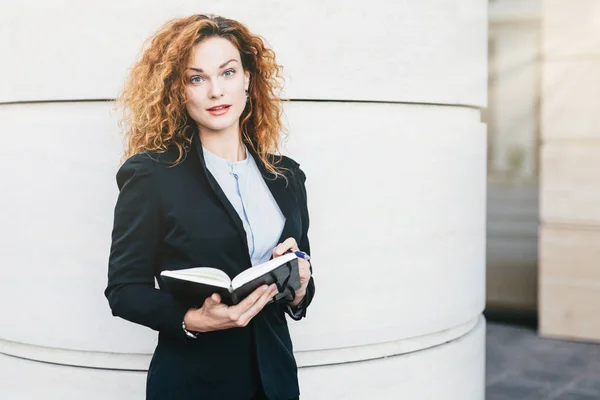 Portrait of businesswoman with curly hair, red painted lips, wearing elegant clothes, writing in her diary book. Good-looking lady model with attractive appearance holding her notebook with pen — Stock Photo, Image