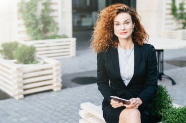 Dalgalı gür saçlı güzel güzel kadın, oturan resmi takım elbise açık kafe, tablet bilgisayar kamera ile kendinden emin bakmak isteyen birisi için beklerken tutan bacak bacak