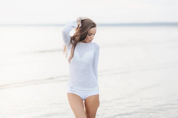 Очаровательная молодая модель в белом свитере и бикини, противостоит красивому морскому пейзажу, задумчиво смотрит вниз, мечтает о чем-то приятном на пляже. Концепция красоты и отдыха — стоковое фото