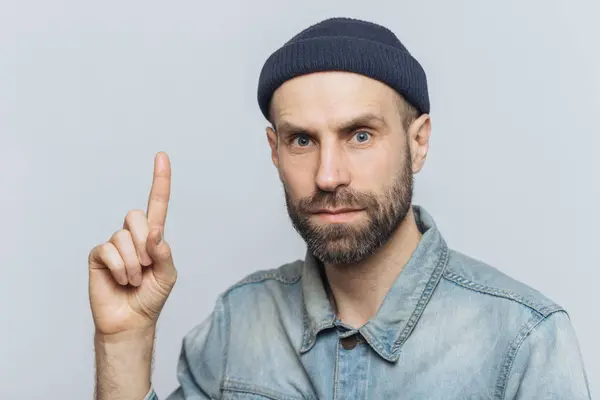 Indoor shot de hombre guapo con aspecto serio, tiene aspecto agradable, barba gruesa con bigote, lleva ropa de moda, levanta figer delantero como se pone brillante idea, aislado sobre fondo gris — Foto de Stock