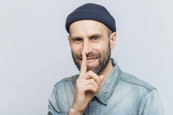 Cheerful attractive male with blue eyes shows silence sign, has satisfied expression, wears fashionable hat and denim jacket, isolated over grey background. Handsome man shows hush sign. Be quiet! — Stock Photo, Image