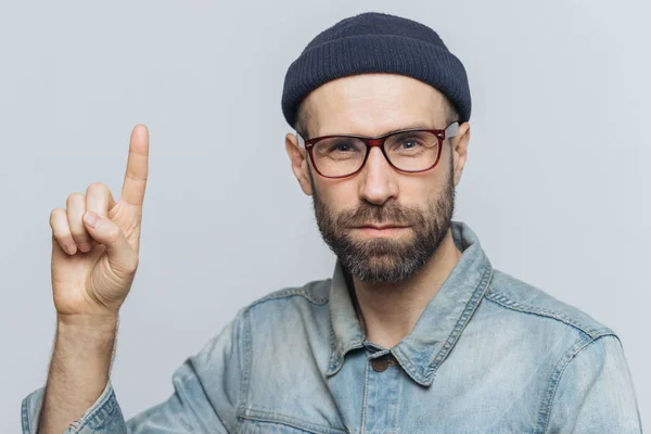 Hombre barbudo serio lleva gafas, sombrero de moda y chaqueta de mezclilla, levanta el dedo delantero como trata de advertirle sobre algo, aislado sobre fondo gris. ¡Sólo escúchame, por favor! Captura de estudio — Foto de Stock