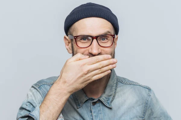 Funny man with attractive look giggles or laughs joyfully, covers mouth with hand, tries to stop feelings, hears anecdote from friend, wears glasses, isolated over grey background. Positive emotions — Stock Photo, Image