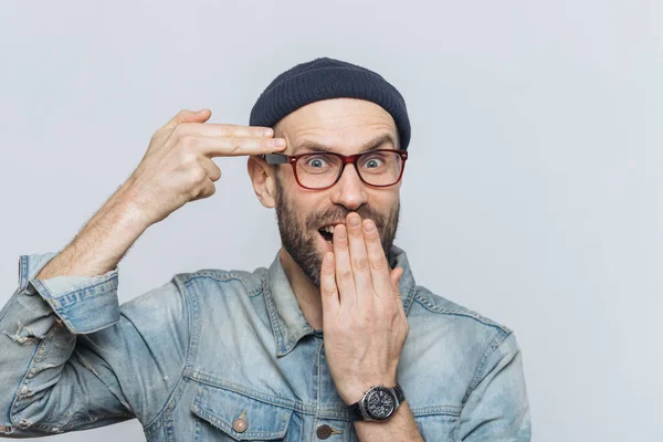 Funny bearded man foolishes indoor, pretends to kiss himself with gun or making suicide gesture, covers mouth with hand, looks joyfully into camera, isolated over grey background. Male foolishes — Stock Photo, Image