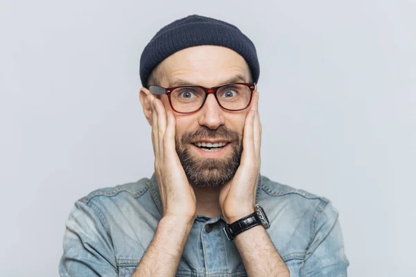 Indoor shot of excited bearded man with blue eyes and cheerful expression, keeps hands on cheeks, dressed in fashionable clothing, isolated over white background. People, facial expressions concept — Stock Photo, Image