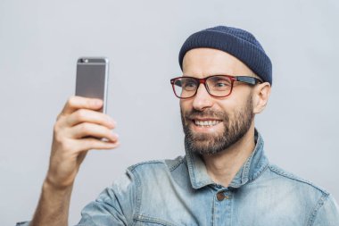 Mutlu orta yaşlı gülümseyen erkek portresi denim ceket, şapka ve gözlük takıyor, selfie, beyaz arka plan üzerinde izole yapmak için akıllı telefon almıştır. İnsanlar, teknoloji ve yaşam tarzı kavramı.