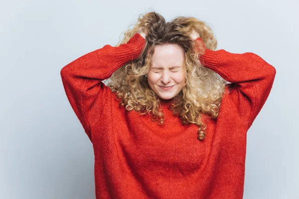 Mujer rubia frustrada estresante arranca el cabello, lamenta hacer mal, expresa emociones negativas, usa suéter rojo suelto, aislado sobre fondo blanco del estudio. Emocional joven hembra interior —  Fotos de Stock