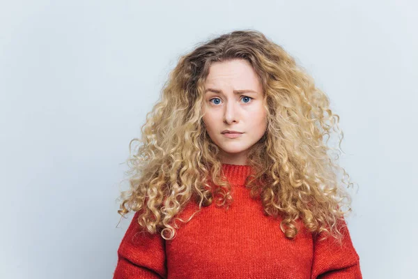 Foto de mujer joven infeliz disgustado con el pelo rubio rizado tupido, frunce el ceño cara en el desconcierto, lleva ropa casual, modelos contra fondo de estudio blanco. Personas y concepto de decepción — Foto de Stock