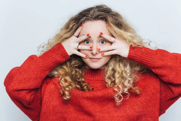 Retrato horizontal de misteriosa hembra de ojos azules con pelo rizado claro, cubre la cara con las manos, vestida con suéter rojo suelto, aislada sobre fondo blanco. Sorpresa y concepto de personas — Foto de Stock