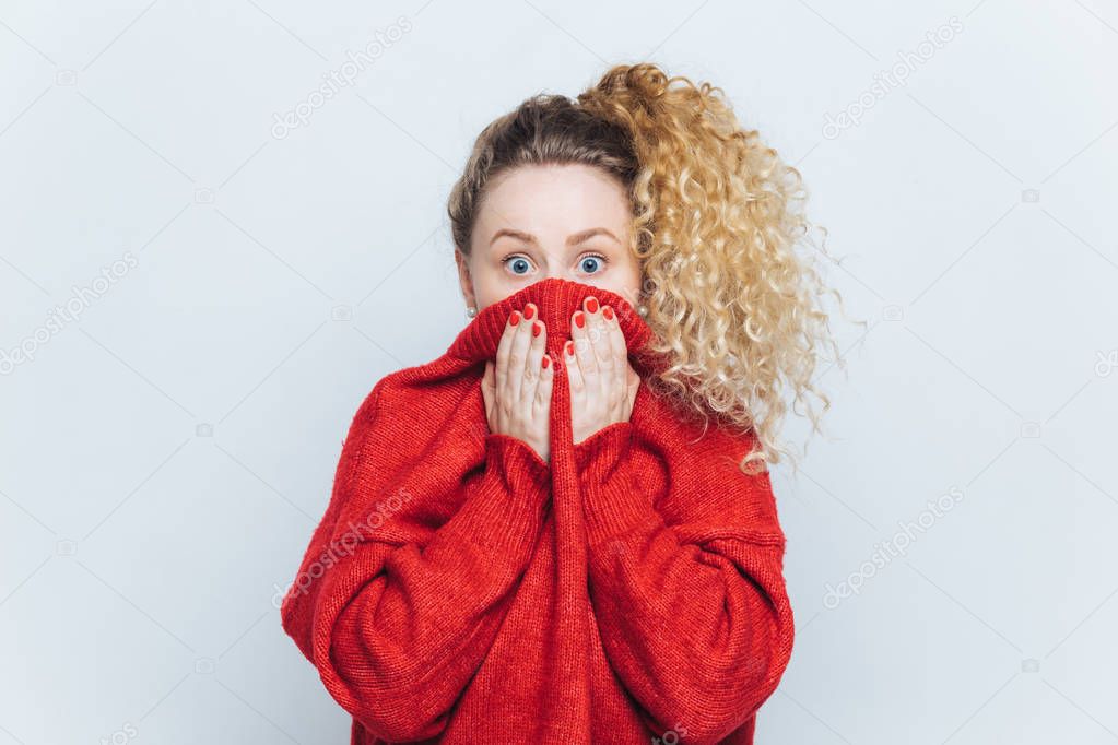 Frightened stunned female sees her phobia, covers face with collar of red sweater, stares at camera with scared look, isolated over white studio background. People, reaction and emotions concept
