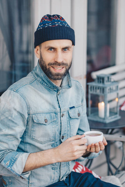 Vertical shot of attractive blue eyed bearded male in stylish clothing, drinks hot tea, looks with pleased expression at camera, spends recreation time at terrace cafe, smiles gently at camera
