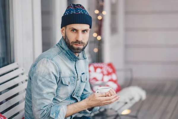 Middle aged bearded male with blue eyes, thick beard and mustache, wears stylish hat and denim jacket, holds cup of hot tea or coffee, has thoughtful expression, thinks about something important