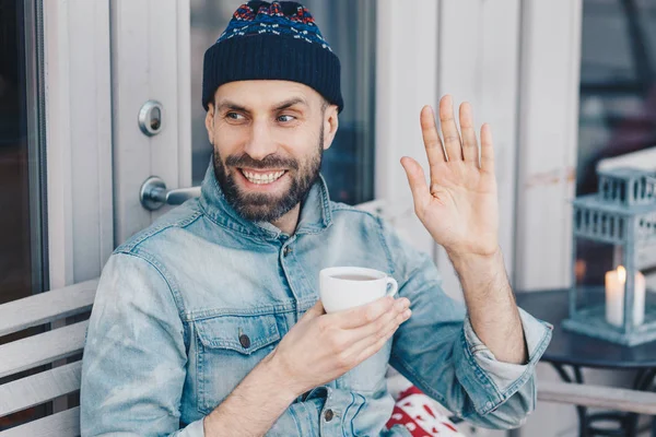 Retrato horizontal de hombre feliz con barba oscura y bigote, olas con la mano, contento de notar amigo, sostiene taza de té, lleva ropa de moda. El hombre saluda a alguien. Hipster chico bebe café — Foto de Stock