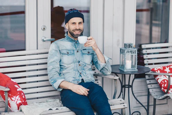Happy blue eyed male with positive look, sits against cozy interior at terrace cafeteria, enjoys aromatic coffee, has spare time after work. Handsome man has beard, looks joyfully at camera.